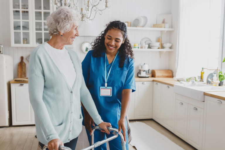Caregiver assisting elderly woman using a walker in a bright kitchen.